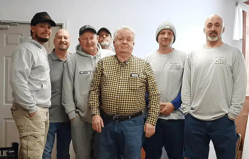 Team of professionals from Mr. Fence It, a chain link fence repair company, posing together in a work setting. The group stands indoors wearing branded work attire, showcasing the skilled crew behind the company’s reliable fence repair services.