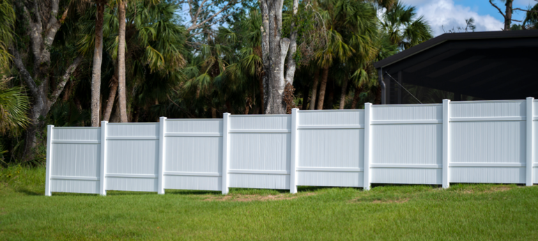 Part of a fence made from white vinyl privacy panels with clean vertical ridges, set against lush green grass and a backdrop of palm trees.