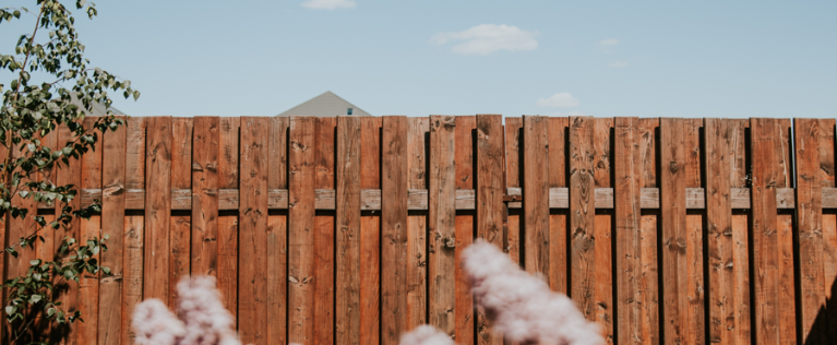 Part of a fence made of reddish stained wood planks with a staggered top, backed by a blue sky and a distant rooftop, framed by soft pink flowering shrubs and a leafy tree branch.