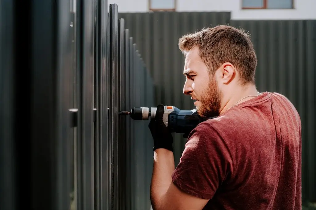 A worker using an electric screwdriver to secure a metal fence panel, demonstrating how to fix a leaning fence by reinforcing the structure.