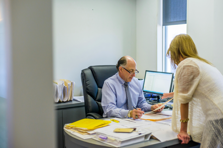 A professional man in business attire sits at a cluttered desk reviewing documents with a woman standing beside him in a modern office setting. The scene suggests a consultation or discussion about property issues, such as determining who is responsible for fence repair between neighbors, with paperwork and legal documents spread out across the desk.