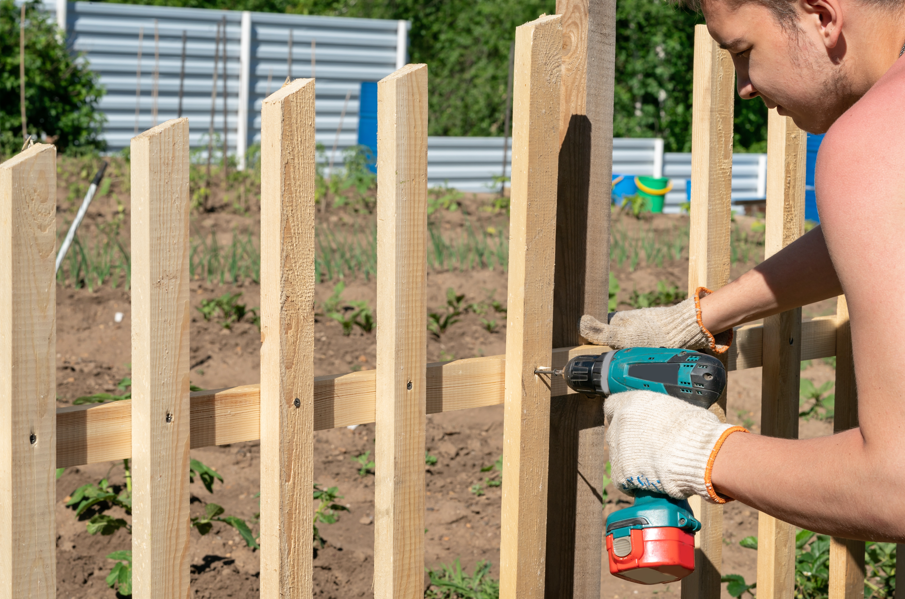 A man in work gloves uses a power drill to secure vertical wooden boards onto horizontal rails, building a fence in a garden plot. This in-progress installation visually captures the labor involved, guiding viewers to consider how much for wood fence installation based on DIY or professional methods.